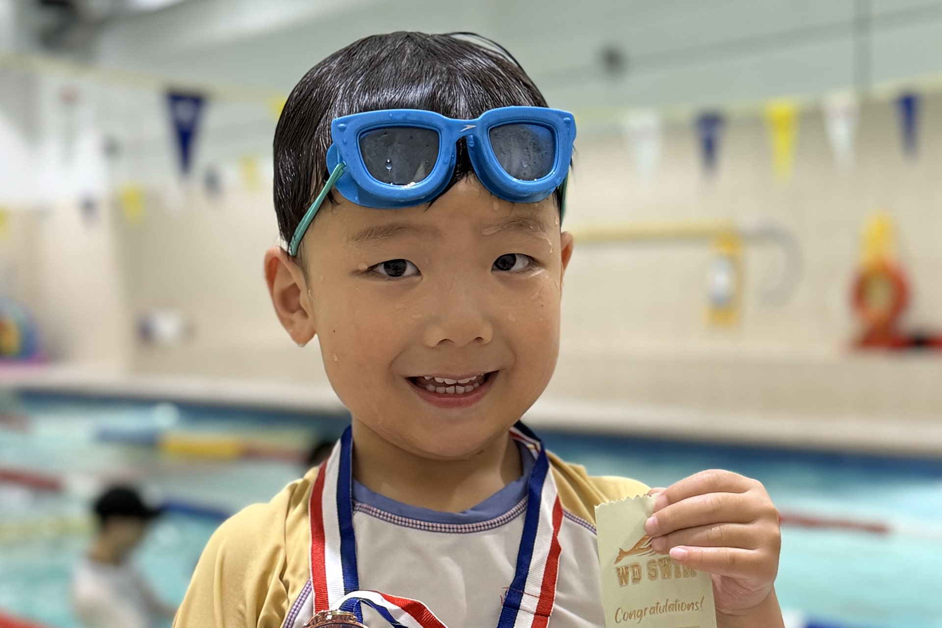 Smiling young swimmer in goggles holding his swimming lesson medal at WD Swim